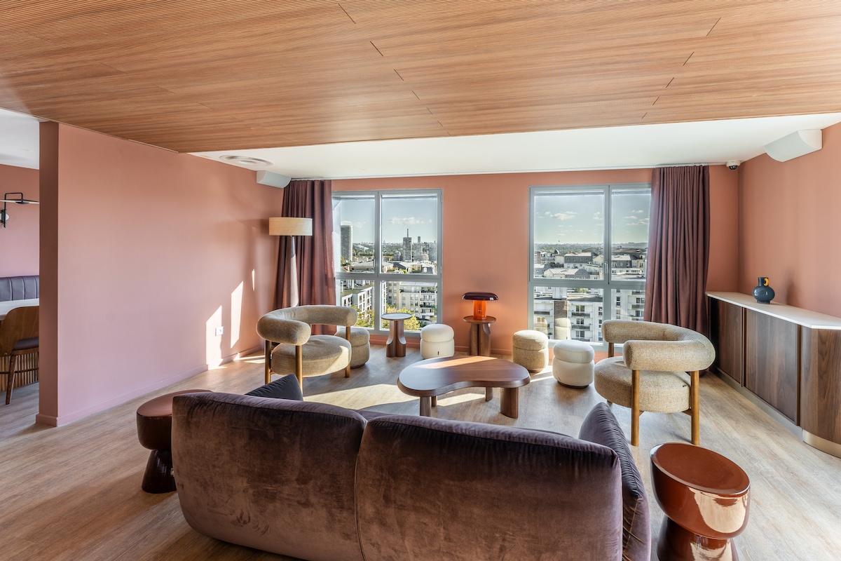 A spacious living area features panoramic windows offering views of the surrounding city skyline. The room is furnished with a velvet sofa and several comfortable armchairs arranged around a wooden coffee table, with soft lighting provided by standing and table lamps.