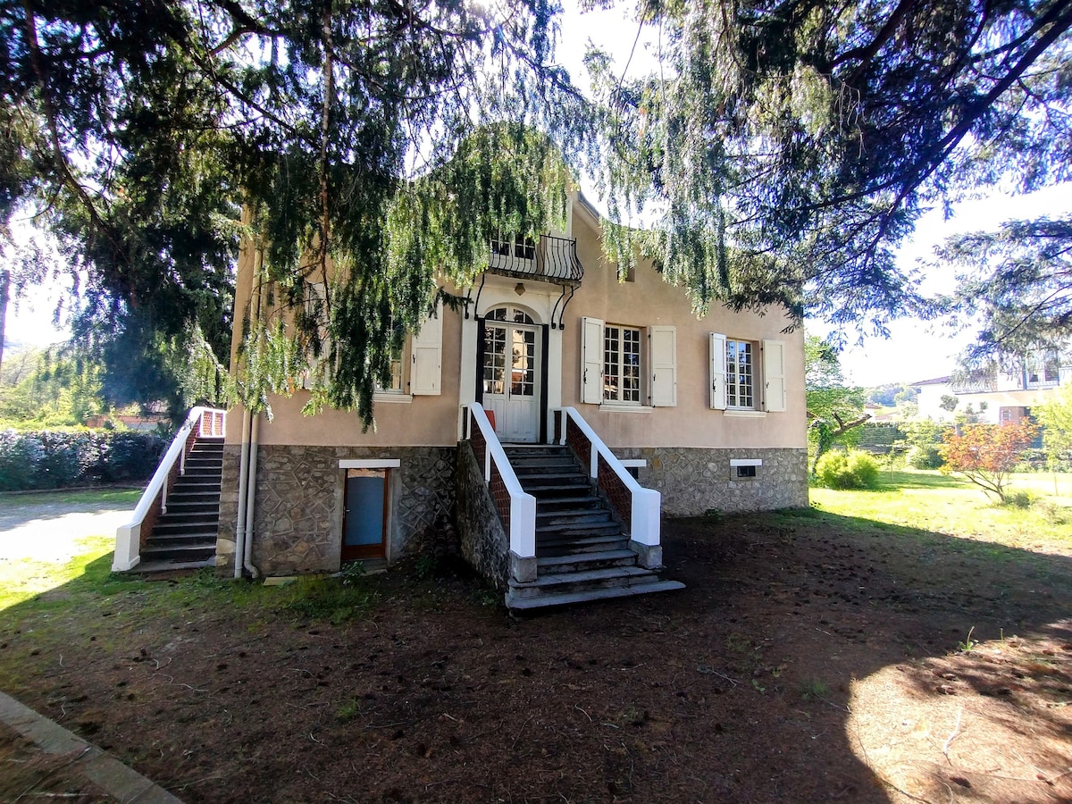 The exterior of a spacious detached property is shown, featuring a charming façade with multiple windows and a balcony. Stone steps lead to the entrance, surrounded by lush greenery and a well-maintained garden area. Sunlight filters through the trees, highlighting the structure.