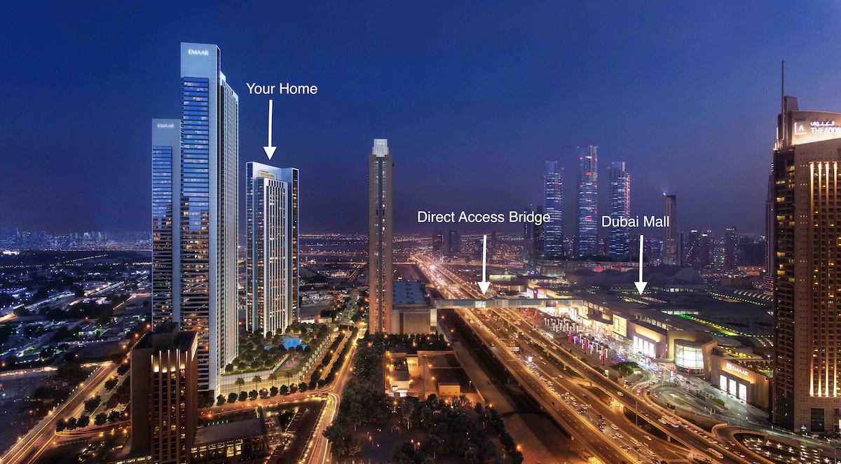 An aerial view captures the apartment complex at night, with modern skyscrapers illuminating the skyline. The direct access bridge leading to Dubai Mall is visible, along with the surrounding vibrant cityscape filled with lights and urban energy.