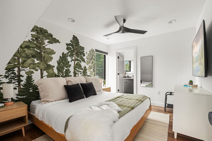 Horná spálňa s manželskou posteľou, pôsobivou lesnou nástennou maľbou, stropným ventilátorom, televízorom a vlastnou kúpeľňou. Útulné útočisko s modernými dizajnovými detailmi a dostatkom prirodzeného svetla.