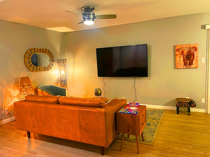 A living area features a full-grain leather sofa facing a large wall-mounted TV. A rustic wooden table beside the sofa holds colorful coasters. Soft lighting from a floor lamp creates a warm ambiance, while a decorative wall piece and mirror add character to the space.