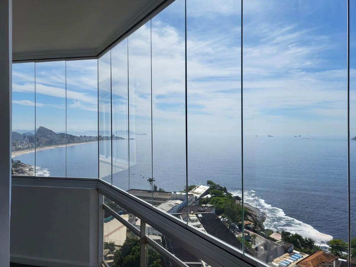 A glass-enclosed balcony offers expansive views of the ocean and surrounding coastline. The tranquility of the water is complemented by distant mountains and a clear sky, creating a serene environment for relaxation. The cityscape and lush greenery can also be observed from this vantage point.