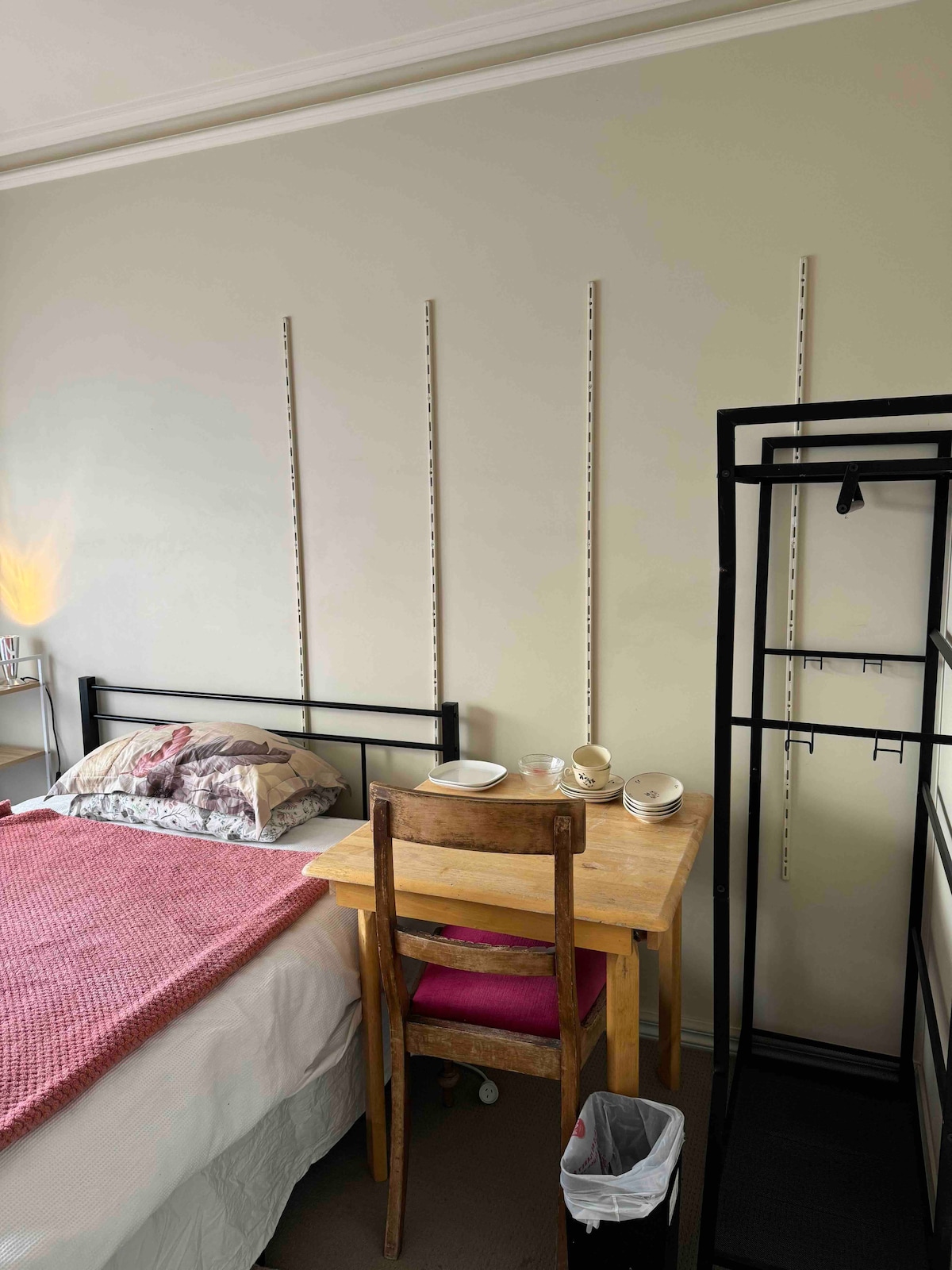 A single room features a bed with a light pink blanket and patterned pillows. A small wooden table with two cups and a chair is positioned next to the bed. A black clothing rack stands nearby, completing the functional layout of the space.