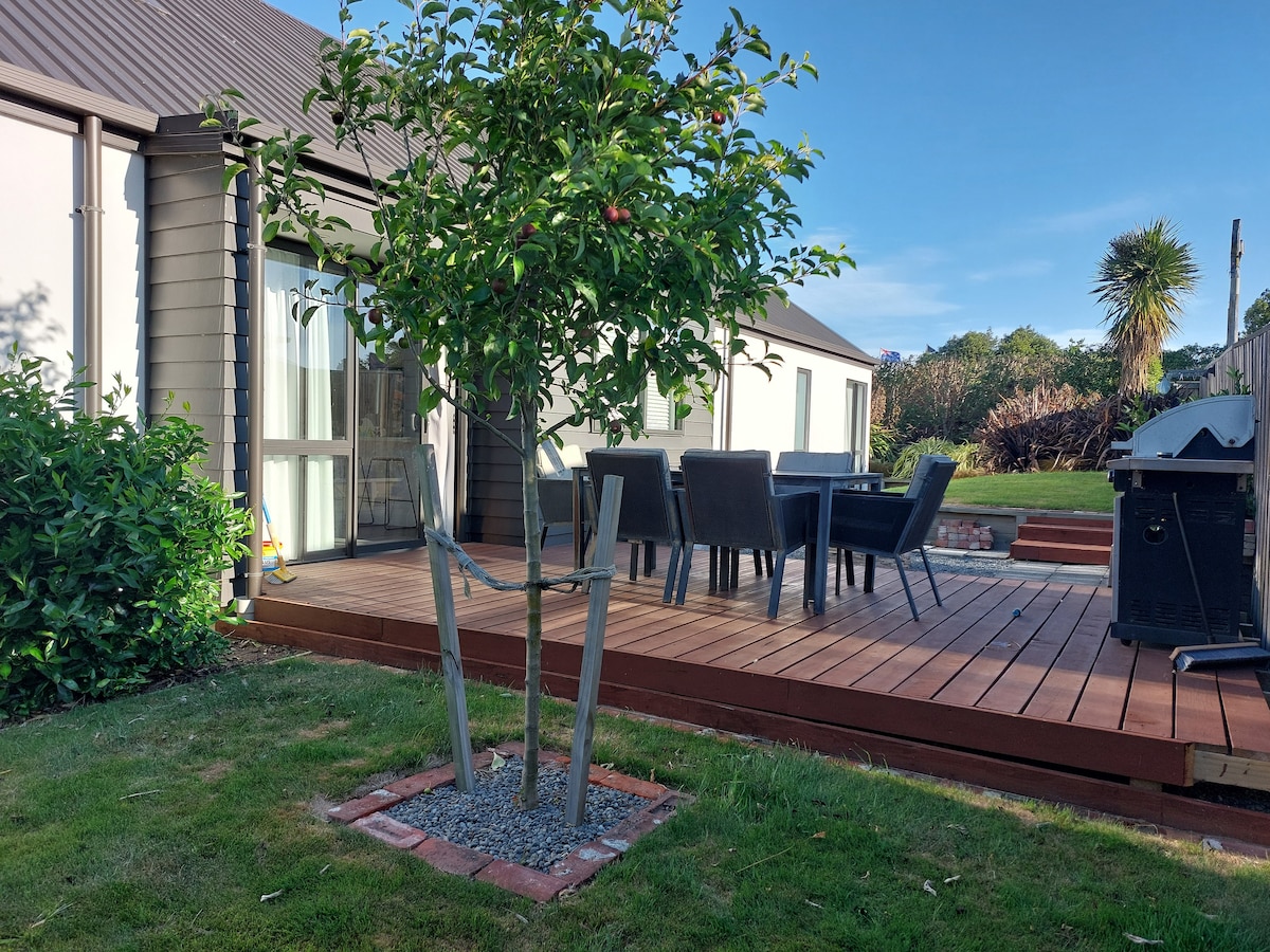 An outdoor deck area features a wooden table surrounded by chairs, offering space for dining and relaxation. A small tree stands adjacent to the deck, framed by well-maintained grass and lush greenery, contributing to a serene outdoor environment.