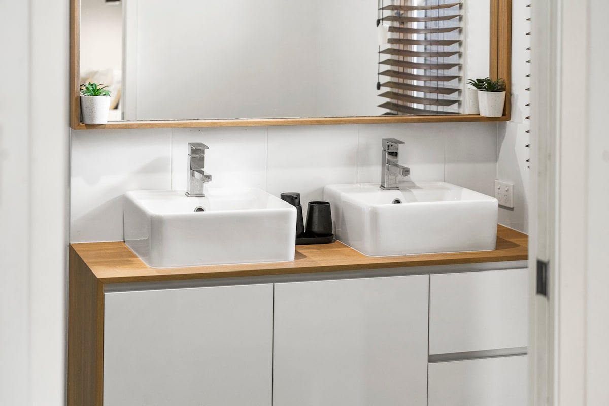A modern bathroom features a double vanity with sleek rectangular sinks positioned neatly on a wooden countertop. Above, a framed mirror reflects the space, while small decorative plants add a touch of greenery. Natural light enters through a shaded window, enhancing the clean, contemporary design.