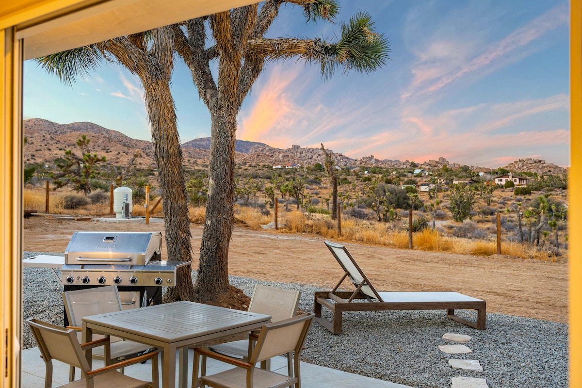 A patio area is visible, featuring a dining table with four chairs alongside a grill. A sun lounger is positioned nearby, and a large Joshua tree is prominent in the scene. The background showcases gently rolling hills and a colorful sunset sky.