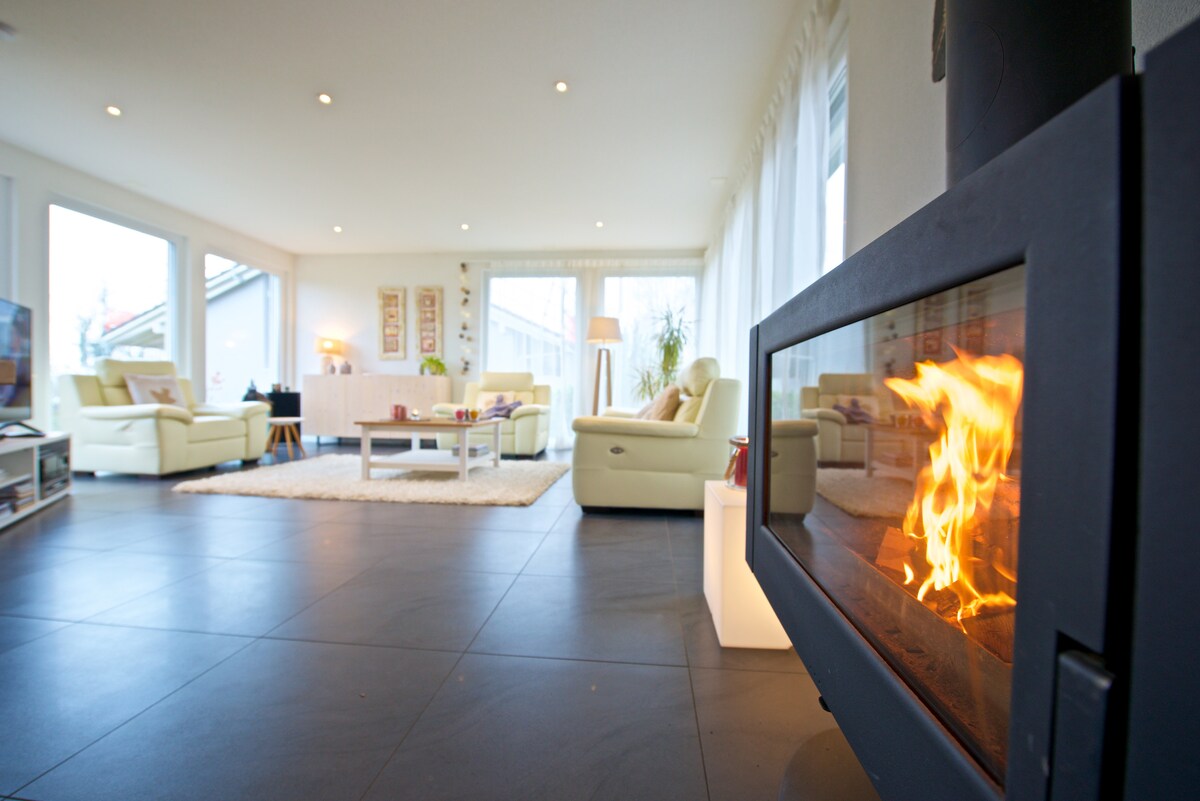 A spacious living area features large windows that allow natural light to fill the room. A modern fireplace stands as a focal point, creating warmth. Comfortable seating arrangements include light-colored chairs and a soft area rug, enhancing the inviting atmosphere of the space.