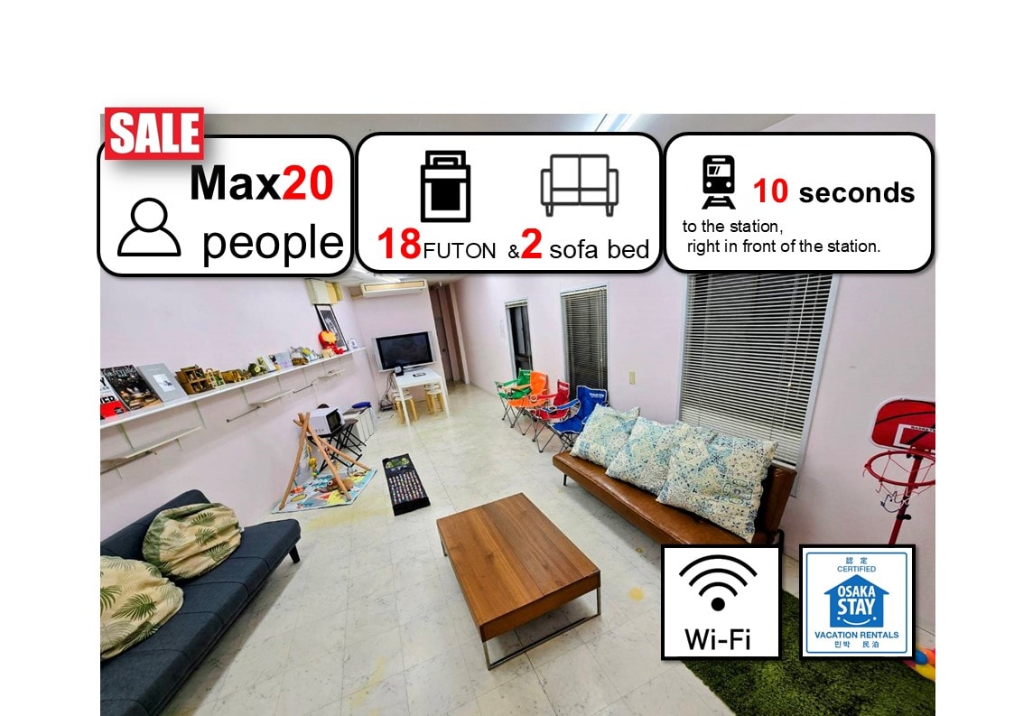 A spacious living area is depicted, featuring a wooden coffee table surrounded by colorful chairs. Two sofa beds and multiple futons are arranged, while a television is mounted on the wall. Shelves display small decorative items, with large windows allowing natural light to fill the space.