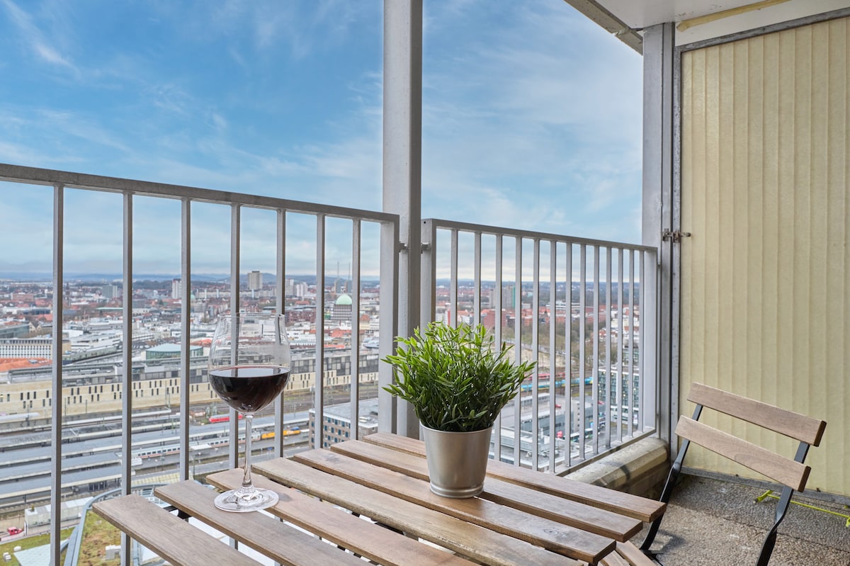 A balcony is shown with a small wooden table and two folding chairs. A glass of red wine is placed on the table next to a potted plant. The view includes a city skyline with buildings stretching into the distance under a clear blue sky.