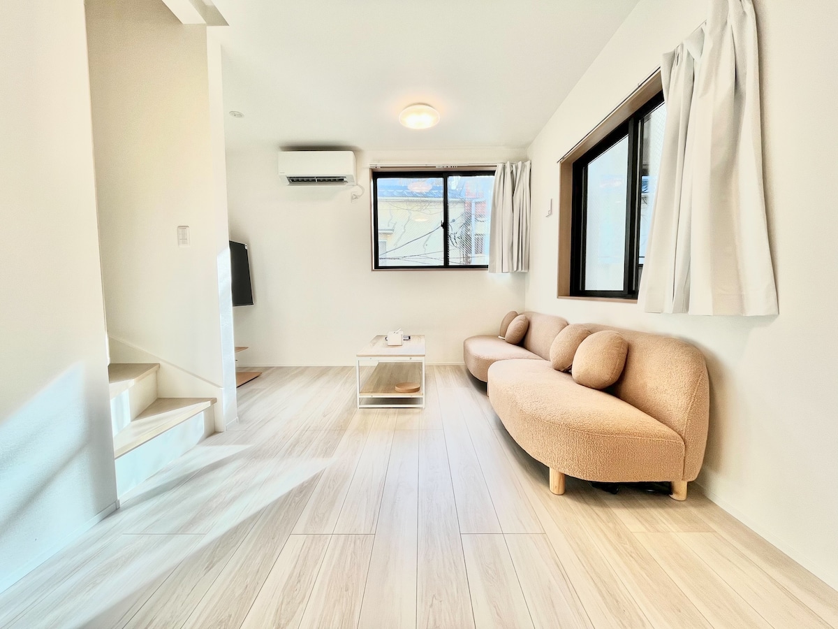 A bright living area is presented with soft natural light filtering through large windows. A rounded, beige sofa is positioned adjacent to a coffee table, creating a welcoming space. Light wood flooring extends throughout, enhancing the open and airy feel of the room.