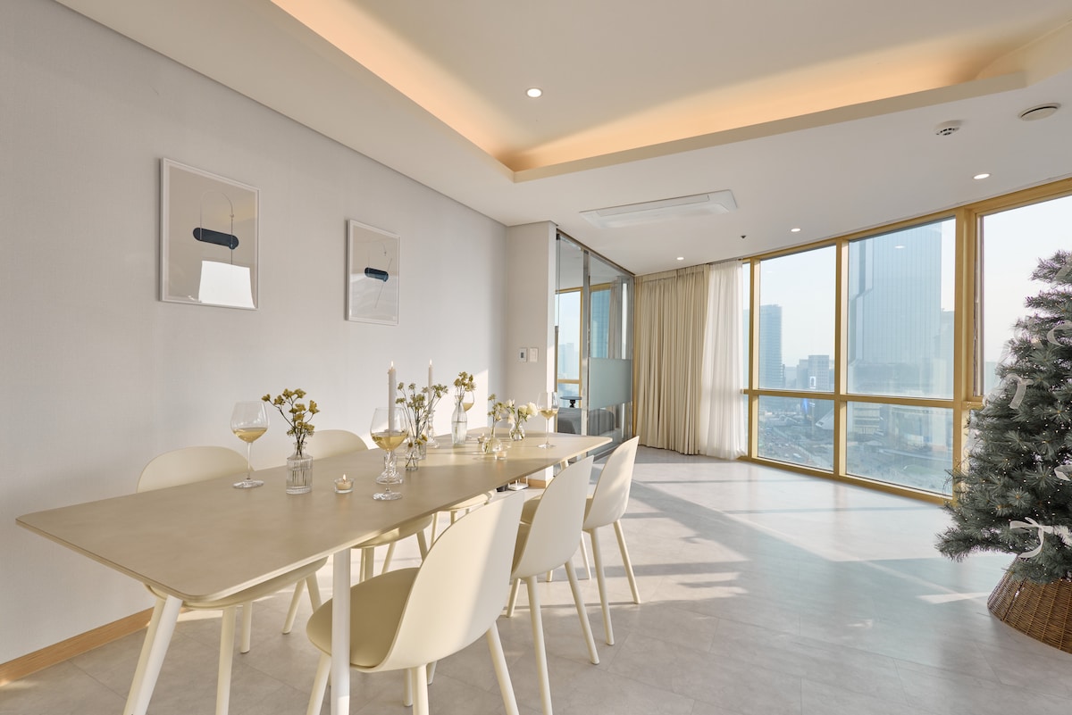 A spacious dining area is featured, furnished with a light-colored rectangular table and six chairs. Natural light floods the room through large windows, highlighting minimalist artwork on the walls. A decorative tree adds a cozy touch to the modern space.