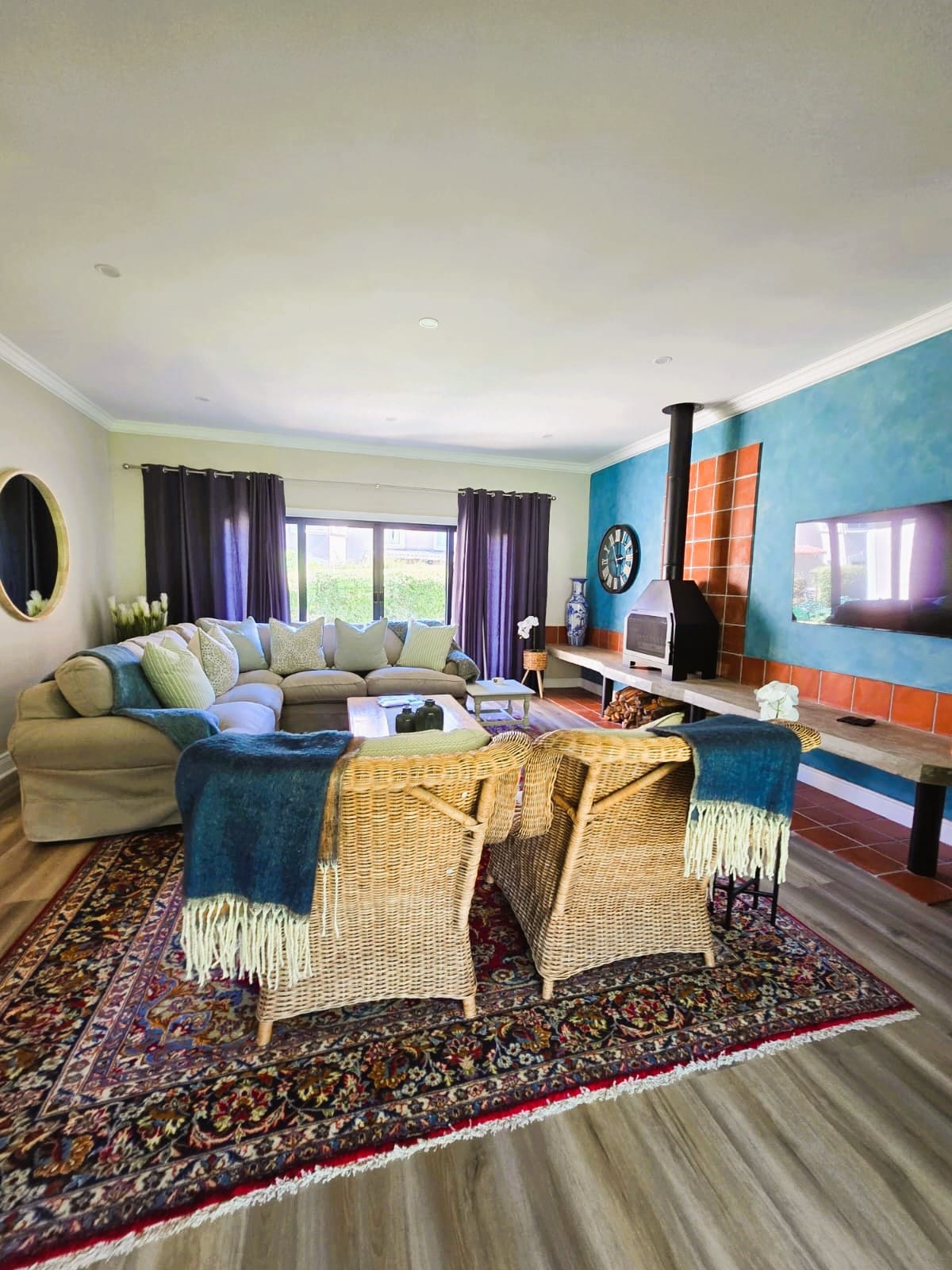 A spacious living area features a large sectional sofa decorated with cushions. Two wicker chairs with throw blankets are positioned near a patterned rug. A fireplace is visible on one side, and windows with dark curtains offer natural light to the room.