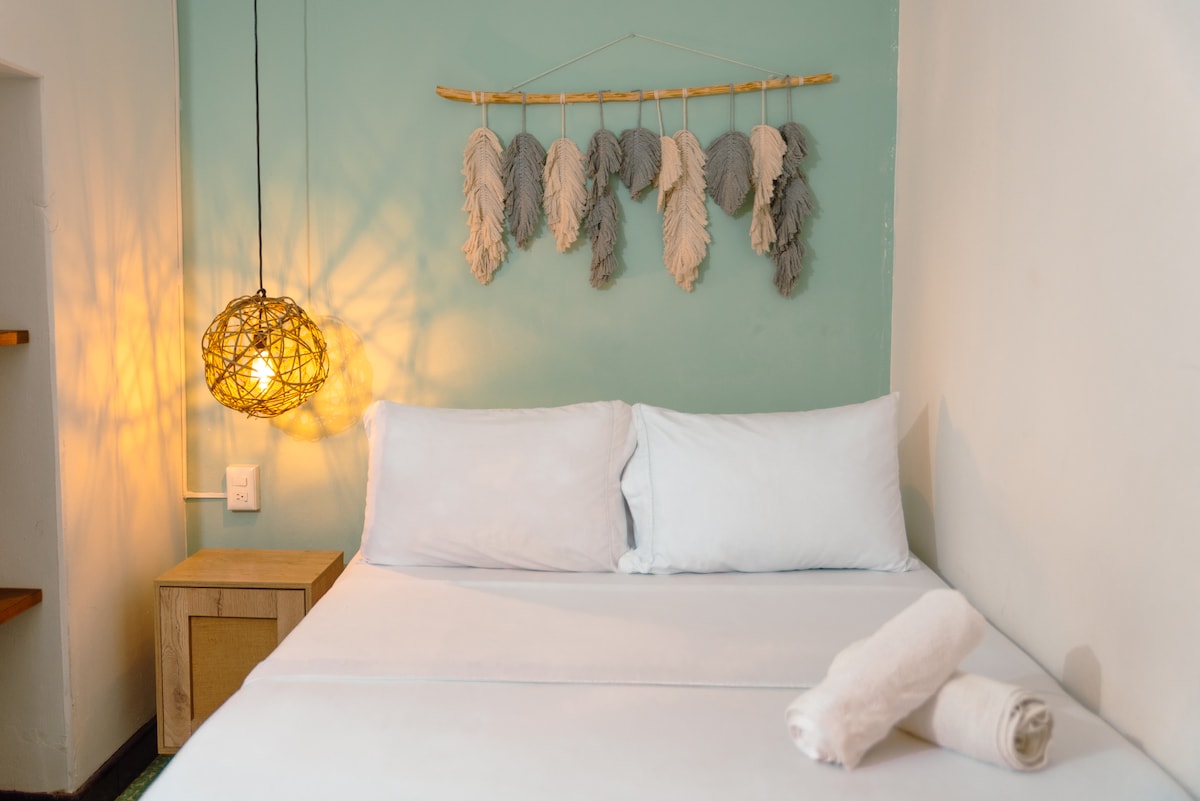 A well-lit sleeping area is featured, showcasing a spacious bed dressed with white linens. A decorative wall hanging with soft hues adds texture above the bed. A natural wood nightstand is positioned beside the bed, complemented by a unique woven light fixture that casts a warm glow.