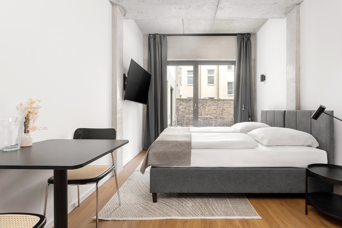 The contemporary bedroom features a spacious bed with crisp white linens and a cozy grey throw. A dining table with modern chairs is positioned nearby. Large windows provide natural light, while dark curtains frame the view. A wall-mounted television is visible across the room.