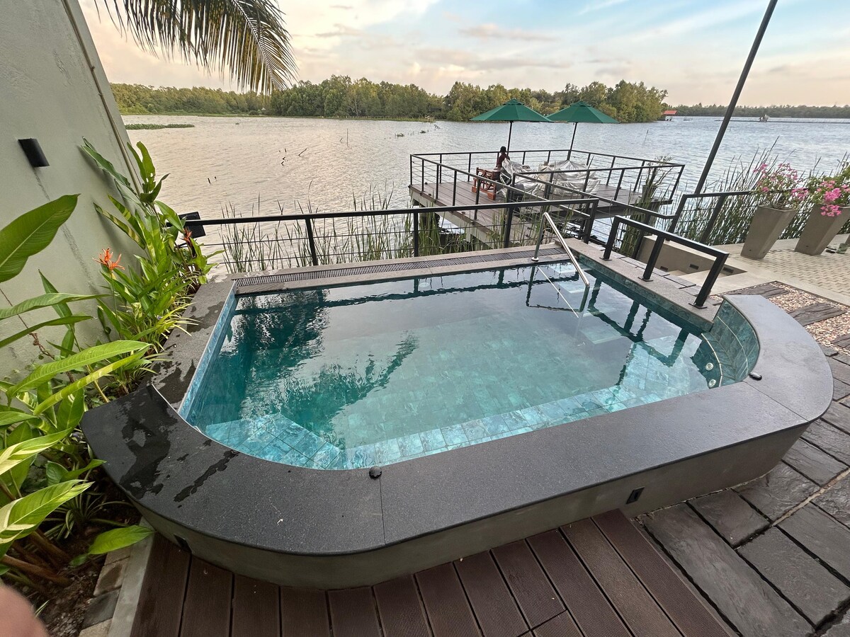 A private swimming pool is situated on a wooden deck, bordered by lush greenery. The pool features a sleek design with steps leading in, and offers serene views of the surrounding water and vegetation.