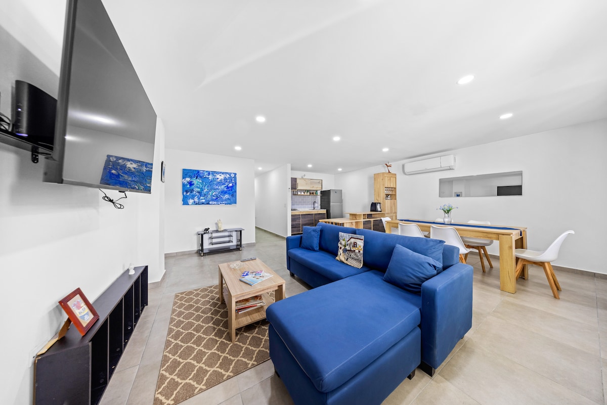A modern living area features a spacious blue sectional sofa arranged around a low coffee table. A large flat-screen TV is mounted on the wall, and an open kitchen with wooden cabinetry is visible in the background. Bright lighting enhances the airy atmosphere.