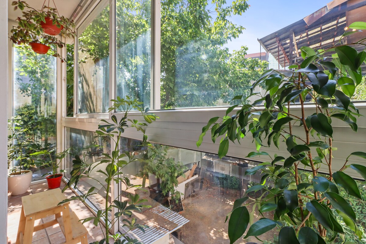 A sunlit balcony is enclosed in glass, showcasing lush greenery and potted plants. Comfortable seating arrangements are visible, inviting relaxation. The surrounding foliage creates a tranquil environment, enhancing the connection to nature just outside the vibrant urban setting.