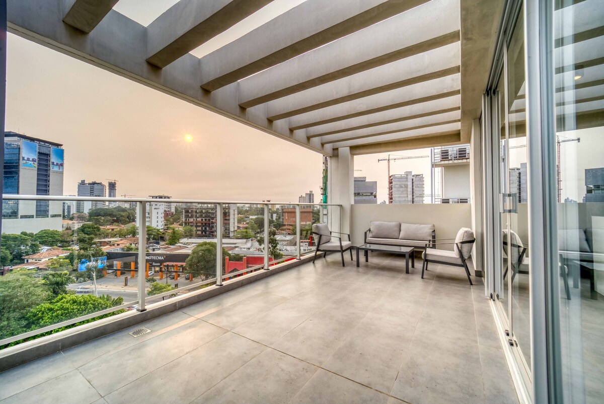 A spacious balcony is featured, offering a view of the surrounding cityscape. Outdoor seating is arranged with comfortable chairs facing the horizon, allowing for a relaxed atmosphere. The protective glass railing enhances safety while providing unobstructed views of the sunset.