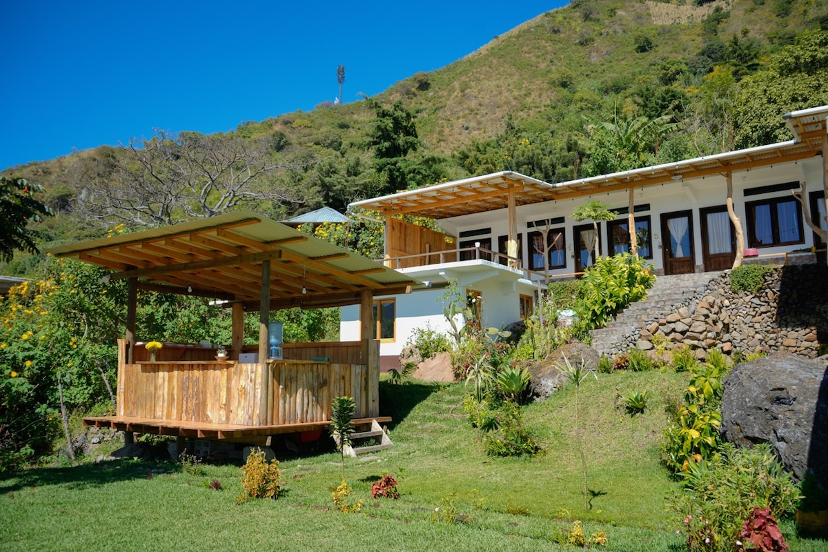 A modern property is nestled against a hillside, featuring a spacious outdoor deck with wooden railings. Lush green grass surrounds the building, while vibrant flowers add color. Nearby, multiple suites with glass doors provide a view of the scenic landscape.