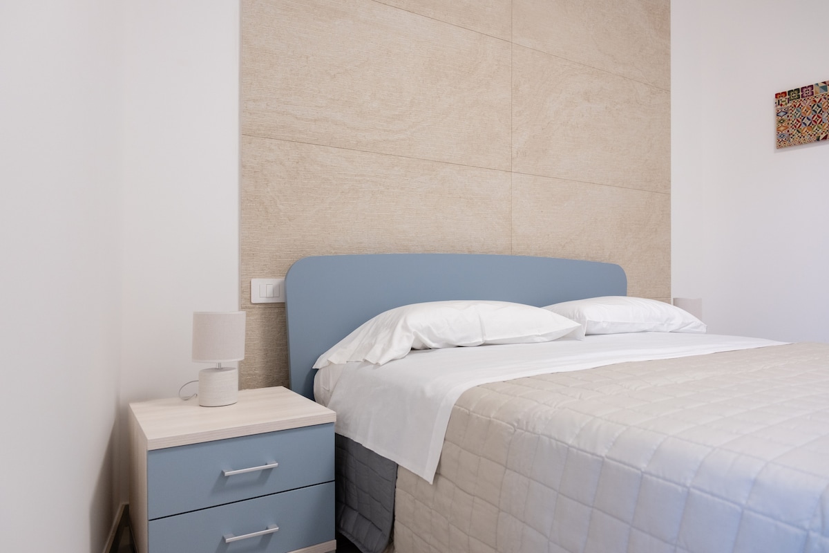 A cozy bedroom features a soft blue headboard above a spacious bed with white linens. A sleek nightstand with a lamp sits beside the bed. The room’s light walls contrast with the textured beige accent wall, creating a soothing environment.