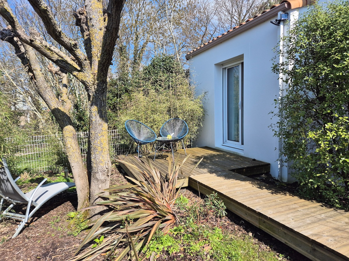A private outdoor space features wooden decking surrounded by greenery. Two chairs are positioned for relaxation, offering a peaceful view of the garden. A large window reveals the interior, allowing for natural light to enter the space.