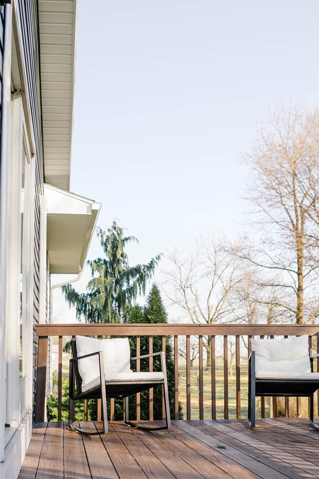A wooden deck equipped with two stylish chairs, each featuring comfortable cushions. The railing provides a clear view of the surrounding trees and open landscape, while a soft sky fills the background, suggesting a calm outdoor space for relaxation.