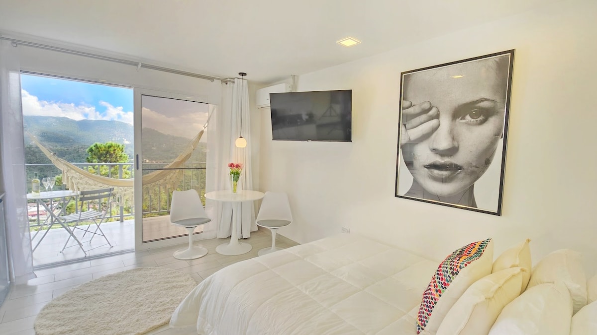 An open-concept mini suite features a cozy queen-size bed and a private terrace showcasing a stunning view. A Smart TV is mounted on the wall, and a round table with two chairs complements the space. Natural light pours in through large glass doors, enhancing the bright atmosphere.