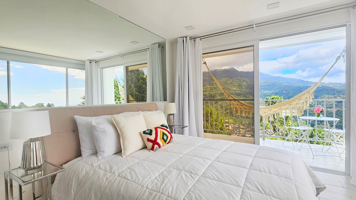 A bright, airy bedroom features a spacious bed adorned with white linens and a decorative throw pillow. Floor-to-ceiling glass doors open to a private terrace with mountain views, a hammock, and a small table with chairs, creating a serene atmosphere.