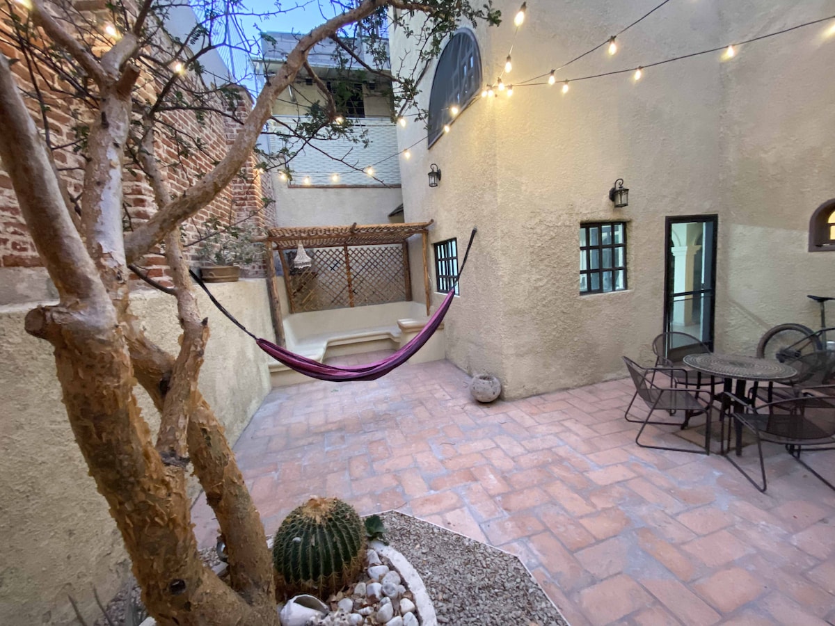 A charming outdoor patio is designed with warm-toned stone tiles and features a hammock suspended between two trees. String lights add a soft glow overhead. A round dining table with chairs sits nearby, surrounded by low-growing plants and decorative gravel.