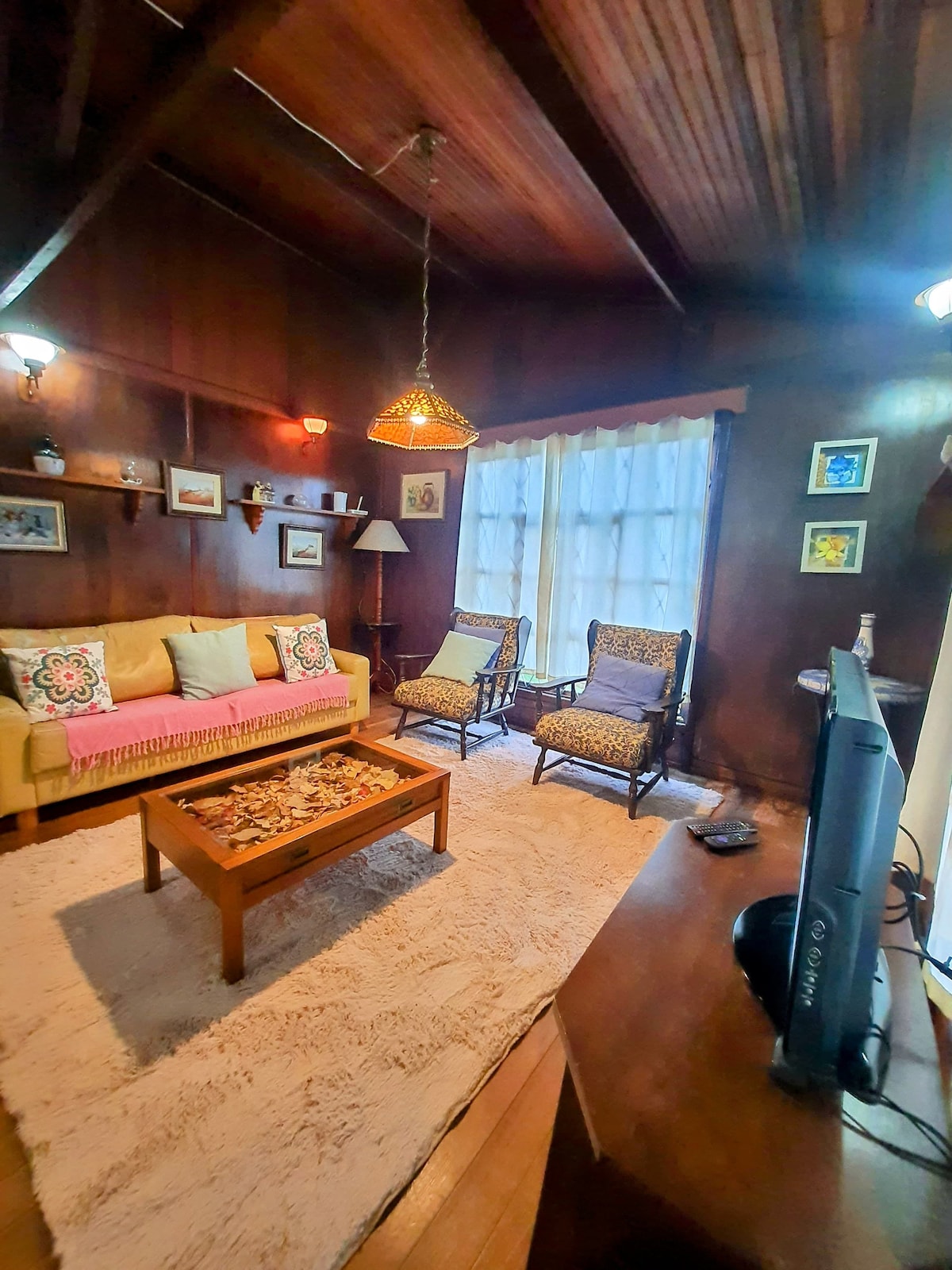 A cozy living area features a light-colored sofa adorned with decorative pillows, complemented by two patterned armchairs. A wooden coffee table sits in the center, displaying a decorative item. Large windows allow natural light to fill the space, revealing warm wooden paneling throughout.