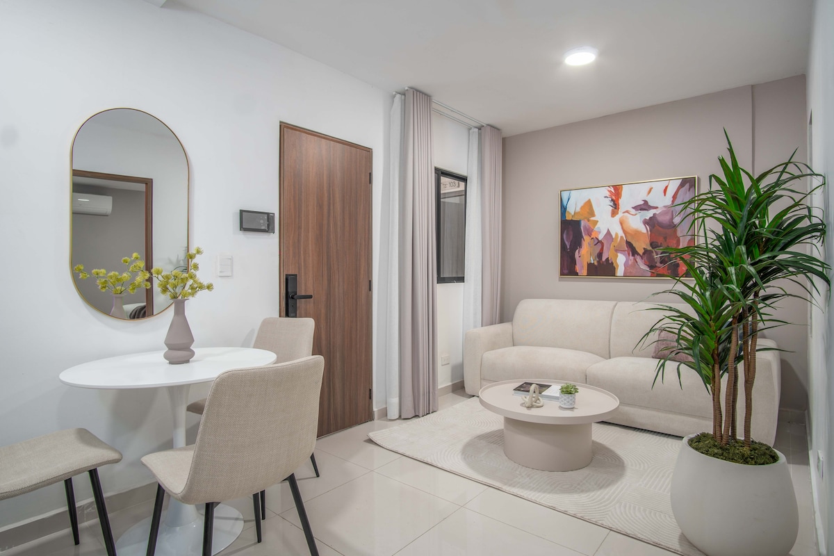 A modern living space featuring a soft, light-colored sofa and a round coffee table. A small dining area with a white table and four beige chairs is visible. A decorative plant adds a touch of greenery, and a large mirror enhances the sense of space.