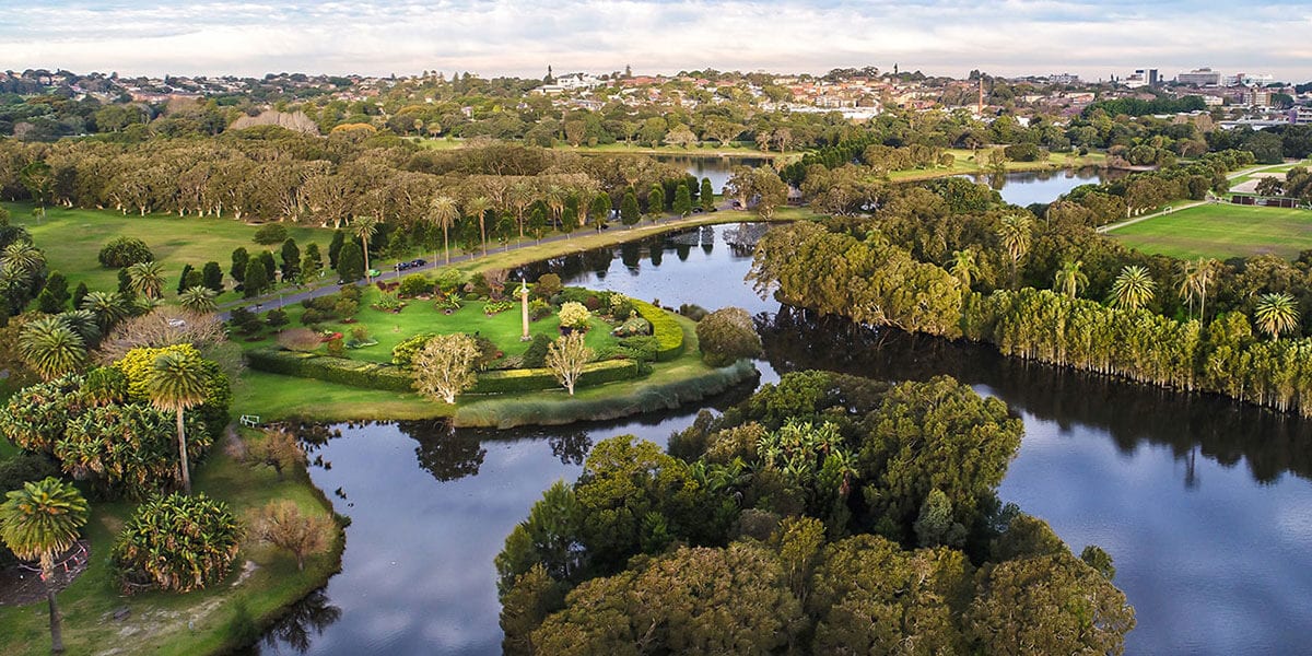 An aerial view showcases lush greenery surrounding tranquil water bodies. Trees and manicured gardens create a serene landscape, with walkways and small bridges visible among the foliage. The distant city skyline can be seen on the horizon, adding depth to the peaceful setting.