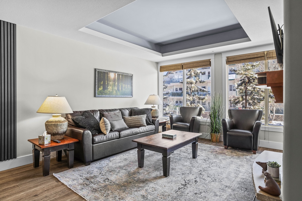The living area showcases a spacious layout with a comfortable sofa and two armchairs. A wooden coffee table is positioned at the center, complemented by soft illumination from two lamps. Large windows provide natural light and views of the surrounding landscape.