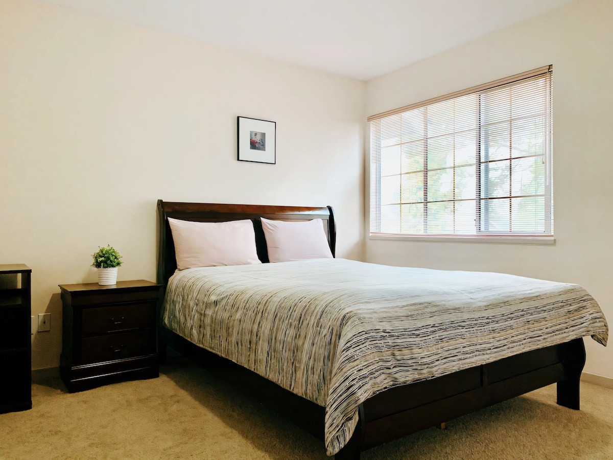 A private bedroom is showcased with a spacious bed dressed in a lightly patterned blanket. Natural light comes through a large window, framed by neatly arranged blinds. A bedside table with a small plant adds a touch of greenery to the calming space.