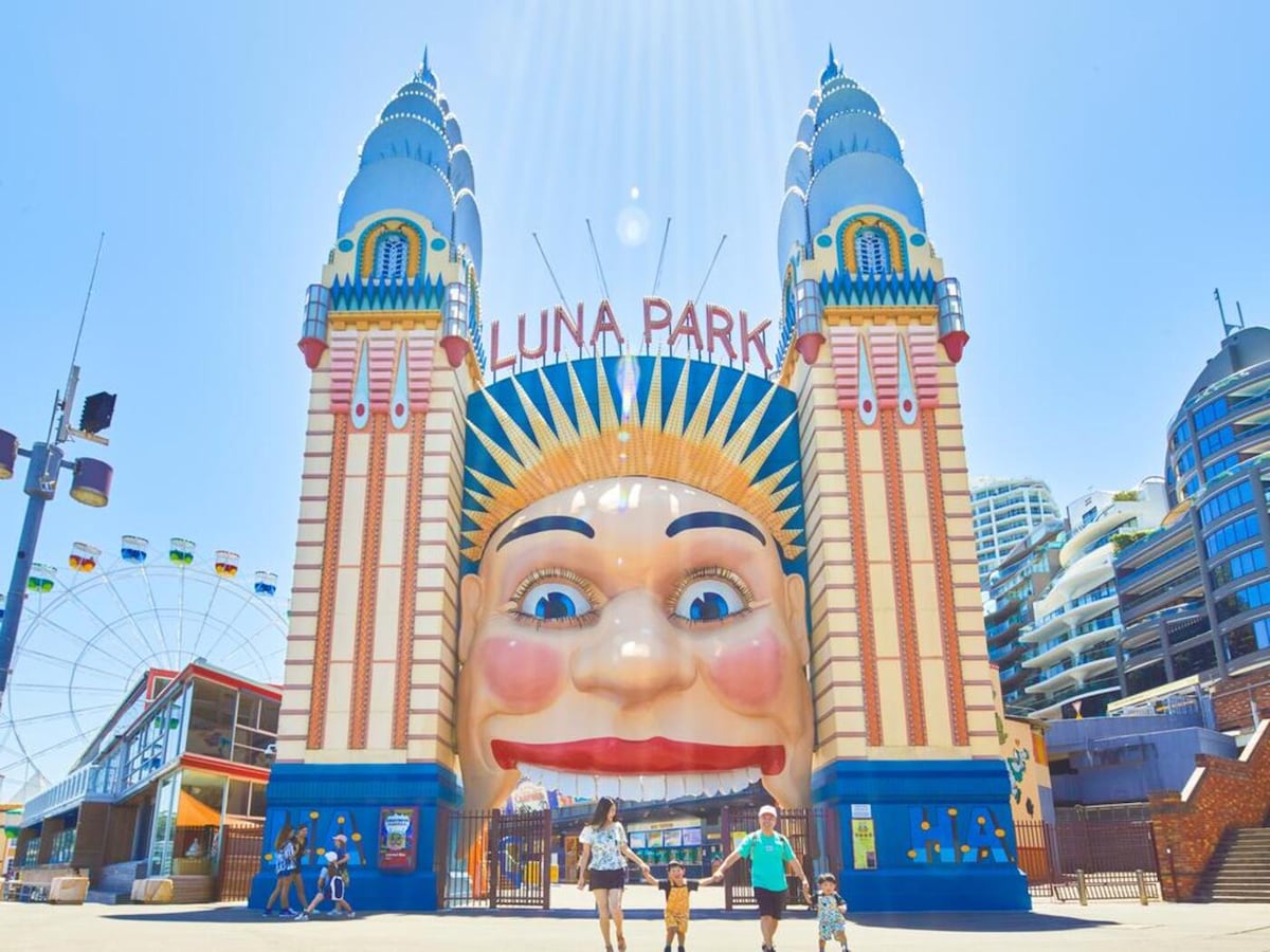 The entrance to Luna Park is displayed, characterized by its vibrant colors and distinctive architectural features. The iconic face structure towers above, framed by blue and cream accents. Visitors can be seen walking towards the entrance, with amusement rides visible in the background.