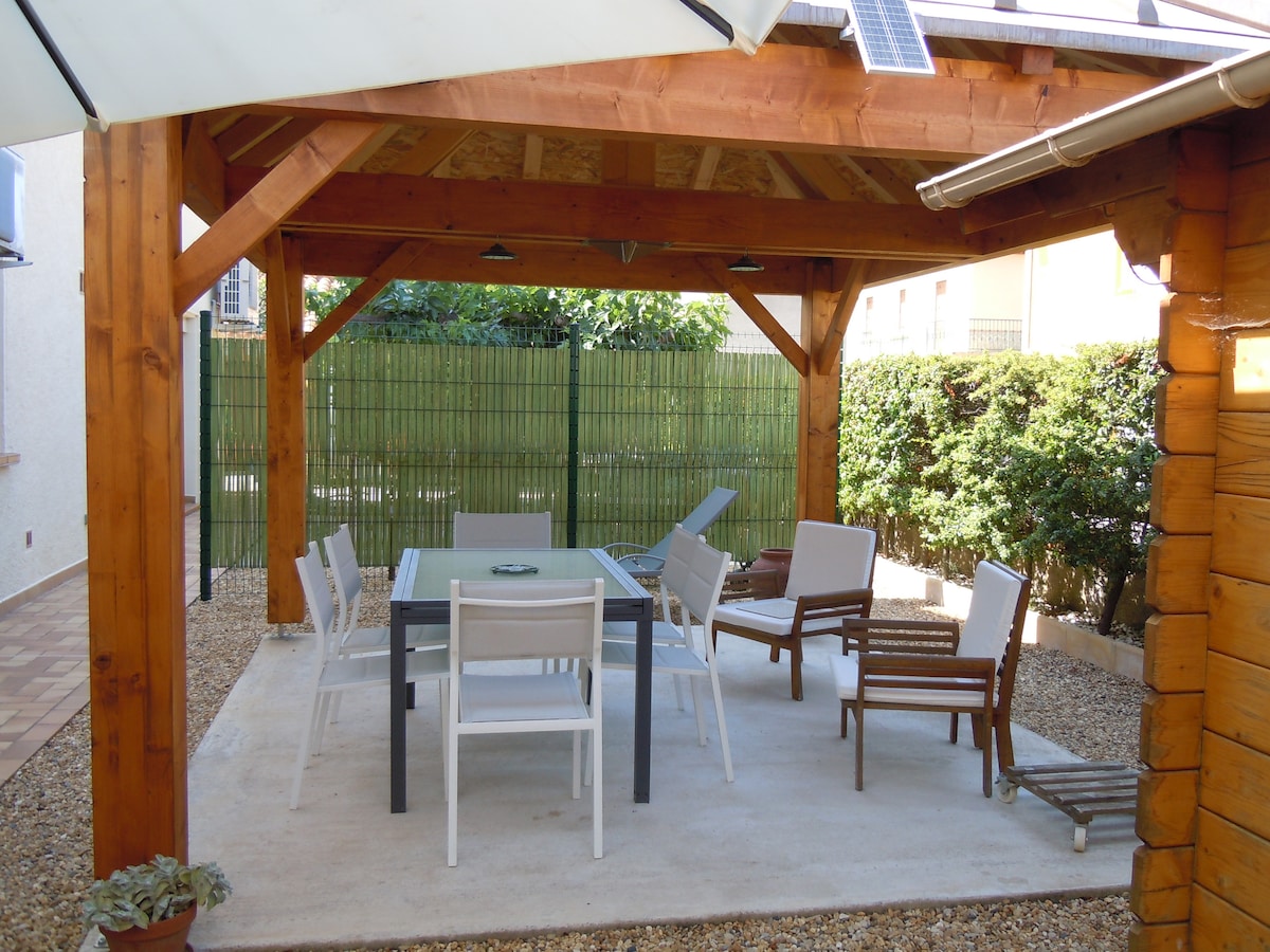 An outdoor terrace is covered by a wooden frame, providing shaded seating with a white and brown color scheme. A rectangular table is surrounded by multiple chairs, set on a gravel floor, with greenery visible in the background, enhancing the space's tranquility.