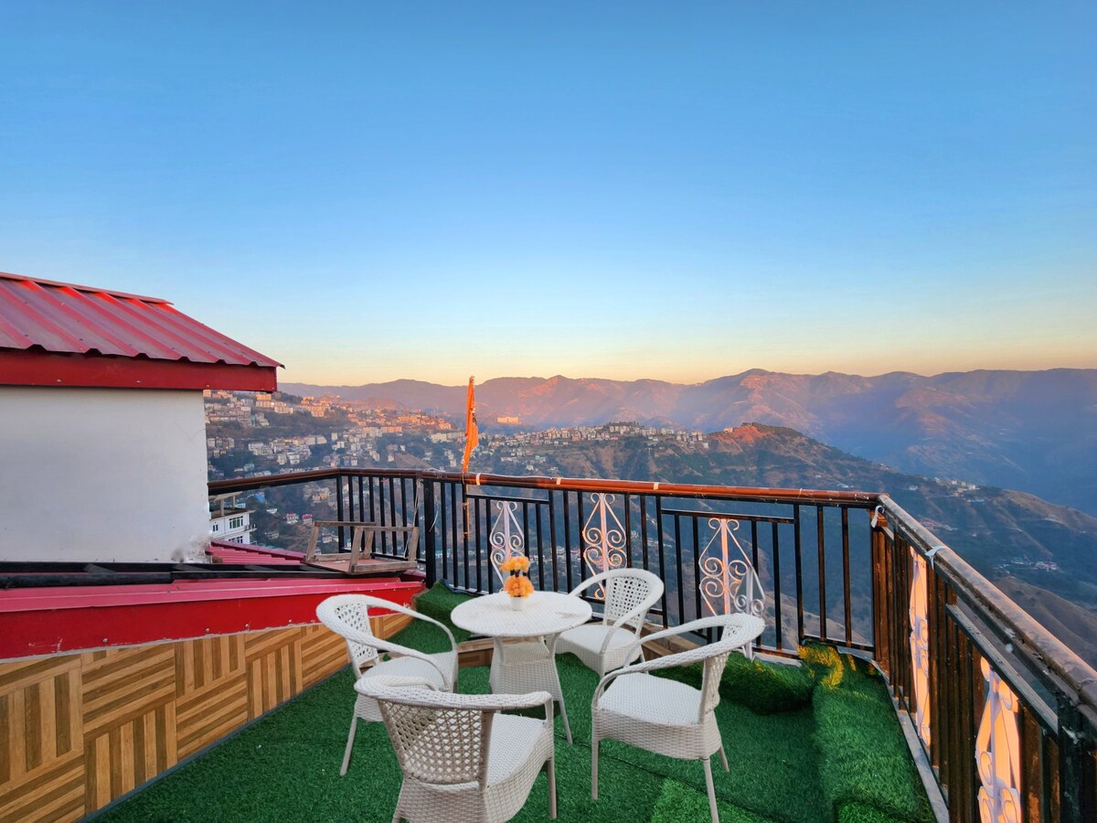 An outdoor terrace is presented with a scenic view of hills in the distance, highlighted by a clear sky. Four white chairs and a small table are arranged on green turf, providing a comfortable space for relaxation and enjoying the landscape.