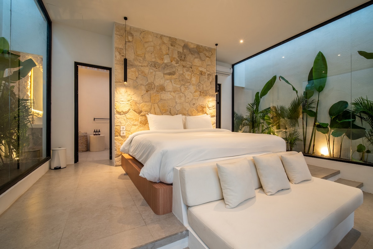 A spacious bedroom features a comfortable bed with crisp white linens. Floor-to-ceiling glass walls showcase lush greenery outside, enhancing the connection with nature. A cozy seating area with neutral cushions complements the design, and a serene ensuite bathroom is visible in the background.