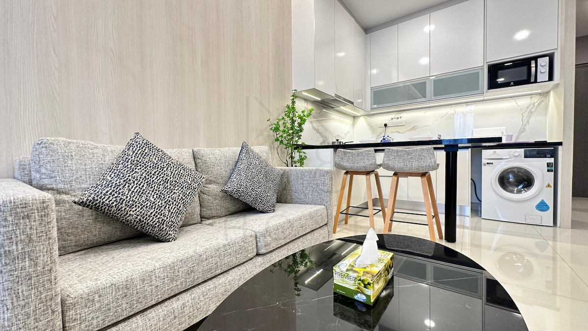 A well-appointed living area features a light gray sofa adorned with decorative cushions. Nearby, a sleek black dining table with wooden legs complements a contemporary kitchen, equipped with modern appliances, including a washing machine. A potted plant adds a touch of greenery to the space.