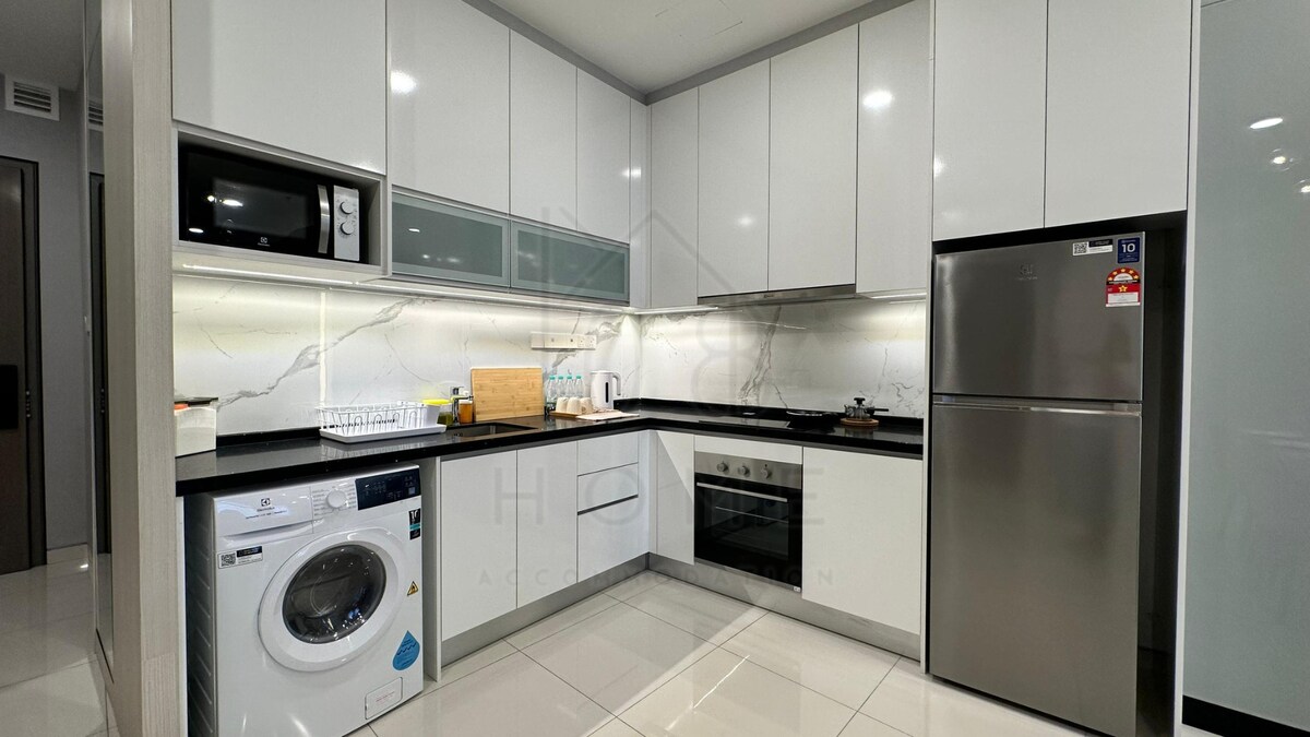 A modern kitchen area features sleek white cabinetry and a marble backsplash. Essential appliances, including a stainless-steel refrigerator, microwave, and washing machine, are present. Ample counter space is available for meal preparation, while soft lighting illuminates the clean lines of the design.