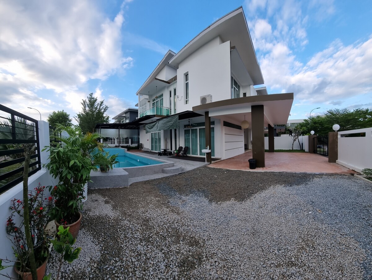 A modern homestay exterior is presented with a swimming pool and a spacious driveway. Minimalist architecture features large windows and a covered entry. Lush greenery surrounds the property, complementing the serene environment under a cloudy sky.