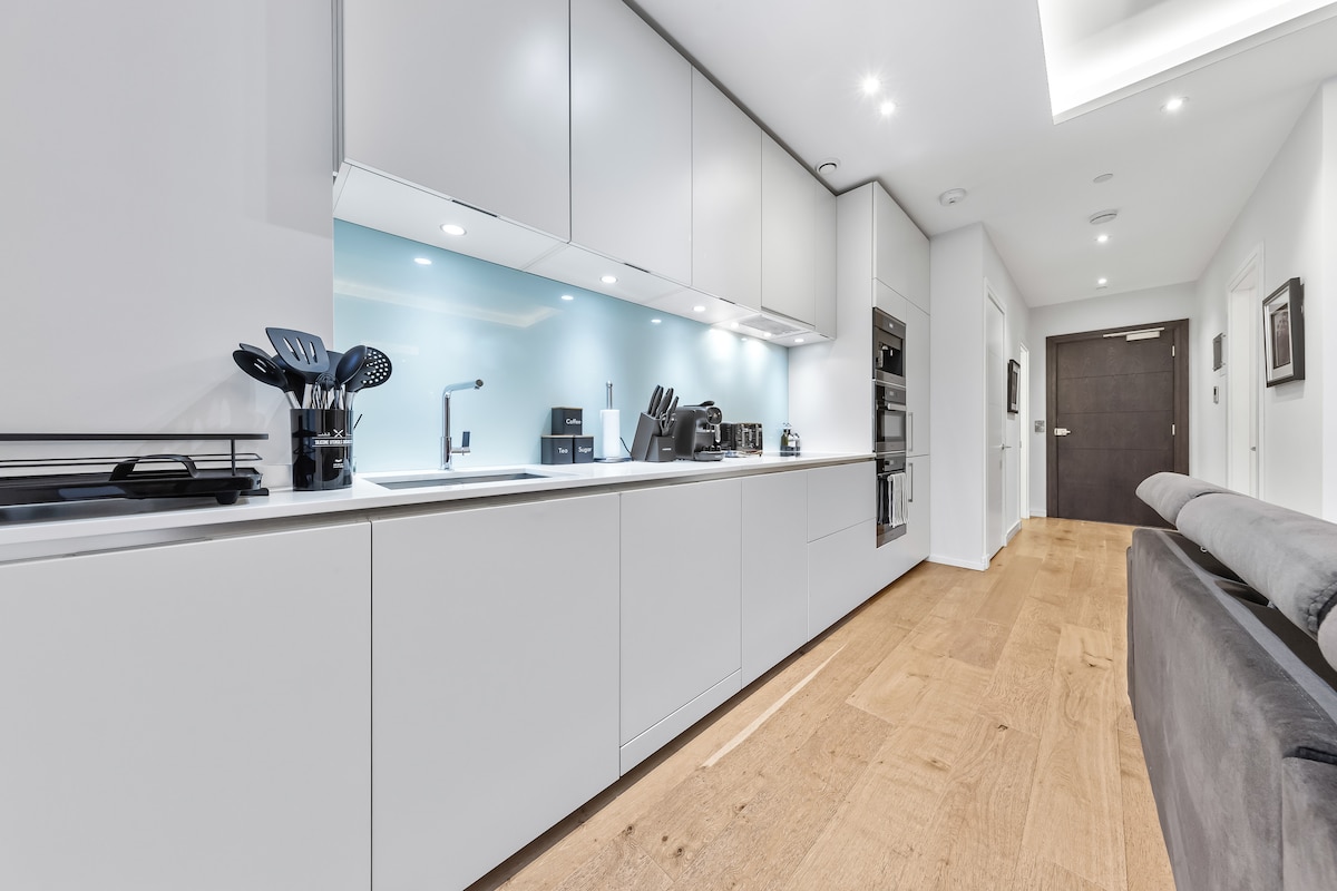 A modern kitchen displays sleek white cabinetry with a minimalist design. Stainless steel appliances, including an oven and a microwave, are visible. A row of cooking utensils is arranged on the counter. Light wood flooring adds warmth to the space, enhancing its contemporary feel.