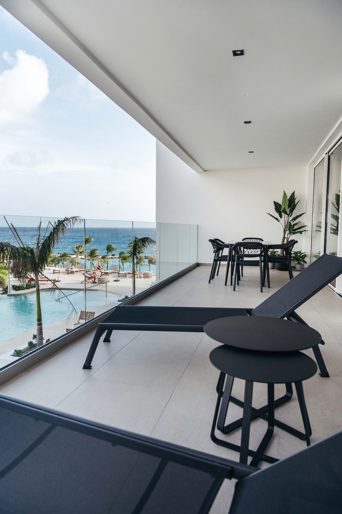 A spacious terrace overlooks the pool and the Caribbean Sea. Minimalistic black lounge chairs and side tables provide space for relaxation. Potted plants are arranged near the glass railing, adding a touch of greenery to the coastal view.