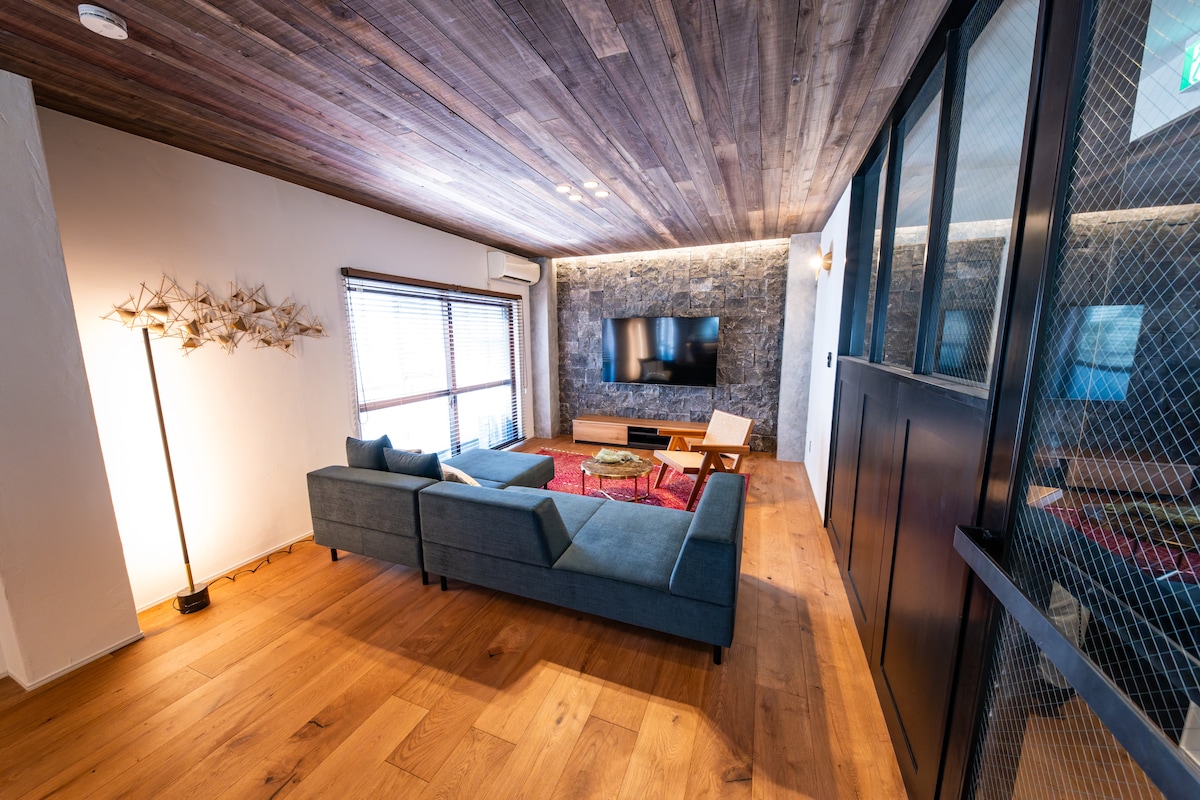 The living area is furnished with a sectional sofa facing a television mounted on a stone wall. Wooden flooring complements the warm tones of the ceiling. Natural light filters through the windows, enhancing the space's inviting feel.