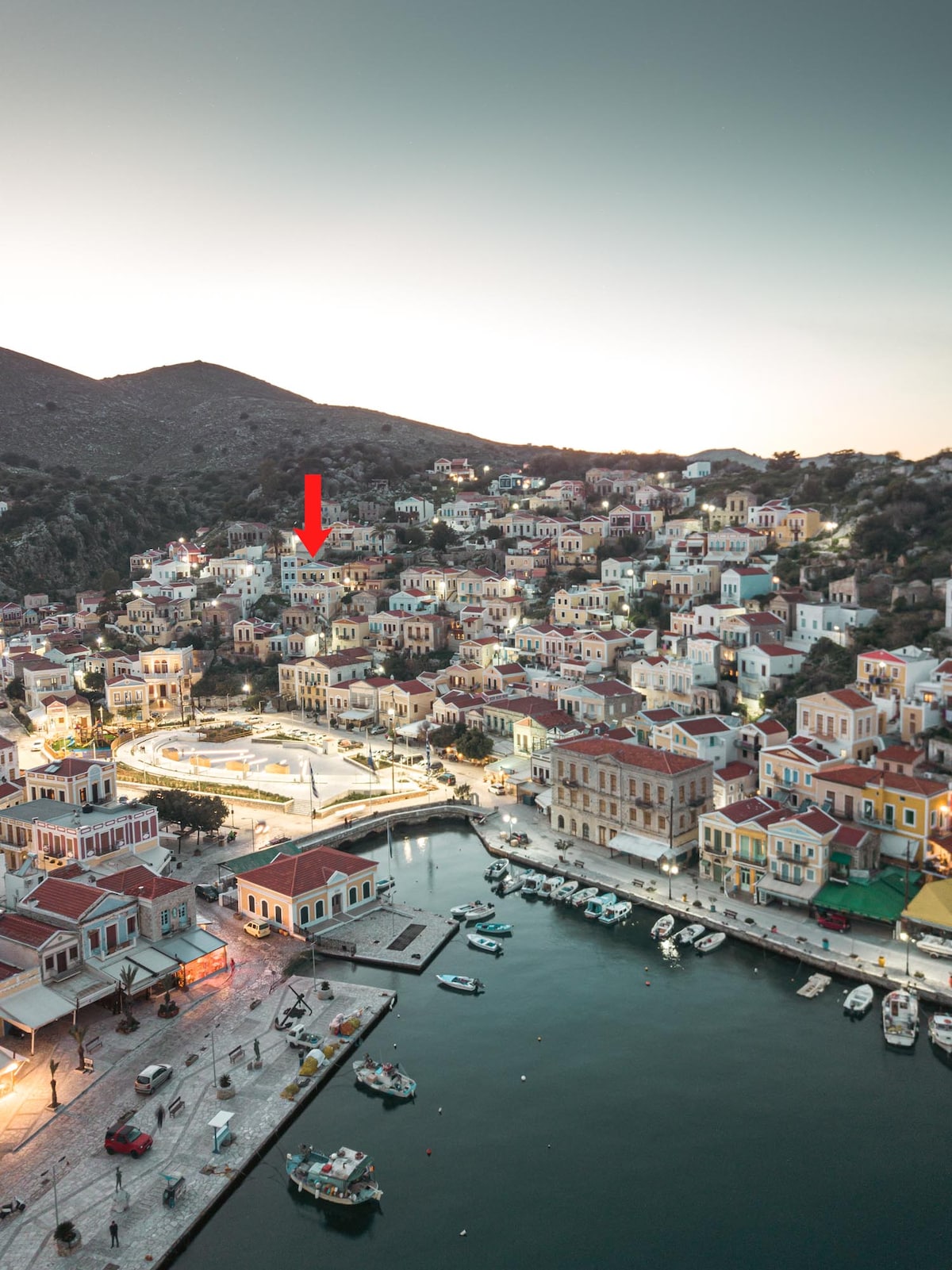 An aerial view of the coastal town of Gialos showcases a collection of traditional buildings with distinctive red-tiled roofs. The Mercury Apartment is indicated by a red arrow, nestled among the hillside residences, while boats are visible in the harbor beneath the soft evening light.