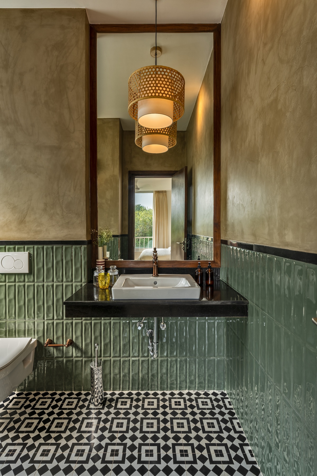 A stylish bathroom features a sleek countertop with a white sink, complemented by dark cabinetry. The walls are adorned with textured finishes and green tiles, while patterned black and white flooring adds character. Natural light filters in through a nearby window, enhancing the space.