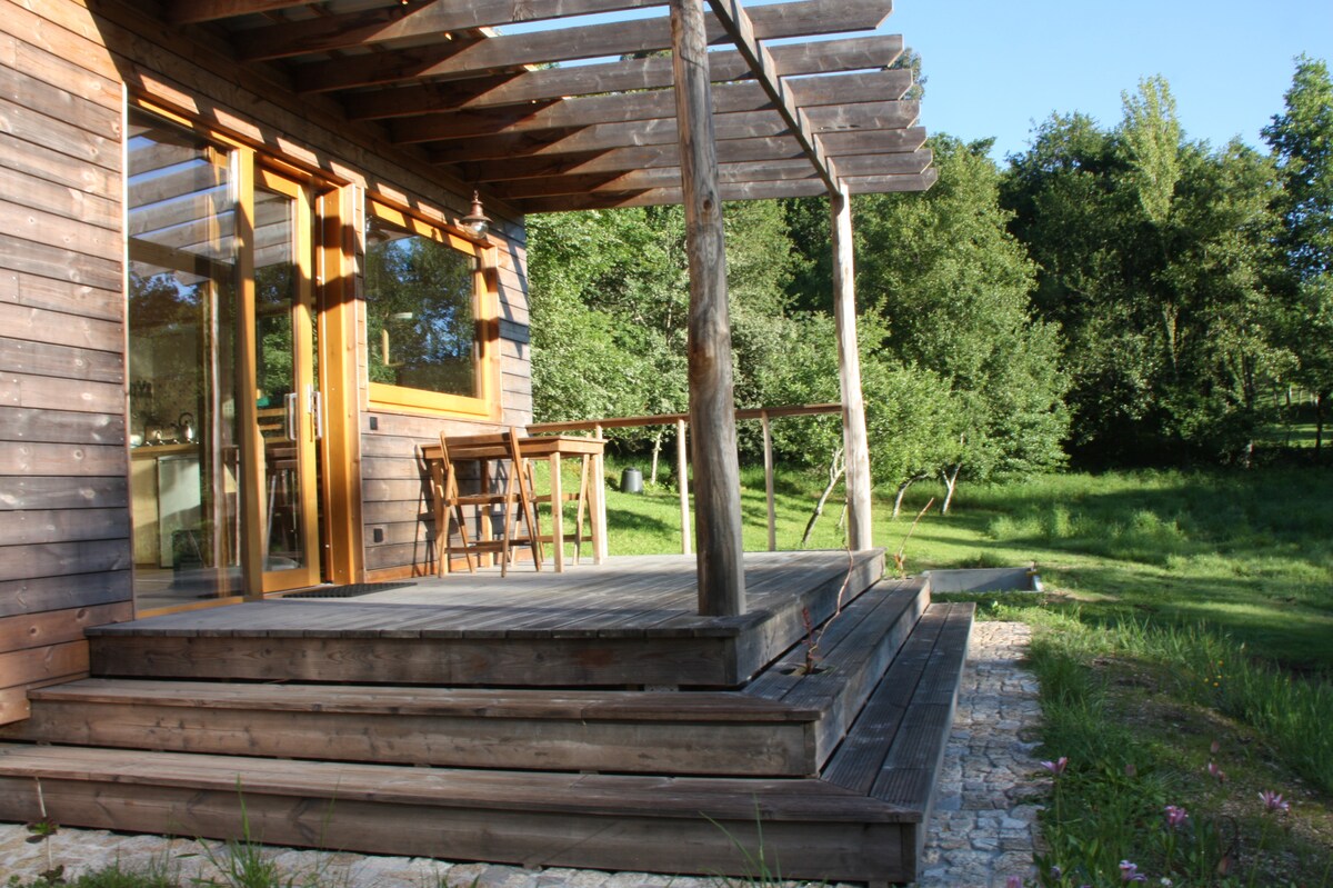 A wooden deck extends from the cabin, featuring a pergola overhead. Outdoor seating can be seen near the entrance. Surrounding greenery adds to the serene environment, while the sunlight casts soft shadows across the patio area.