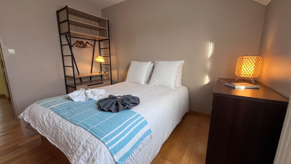 A cozy bedroom features a spacious bed dressed in white linens with a light blue throw. A bedside table is accompanied by a warm lamp, and a modern bookshelf holds folded towels. Natural light filters in through the nearby window, creating a calm environment.