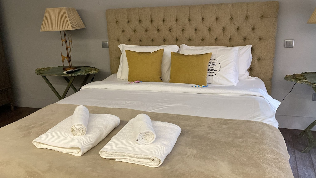 A spacious bed is dressed in crisp white linens, with two yellow decorative pillows placed against a tufted headboard. Two folded towels rest on a muted beige bedspread, and bedside tables with lamps are positioned on either side of the bed.