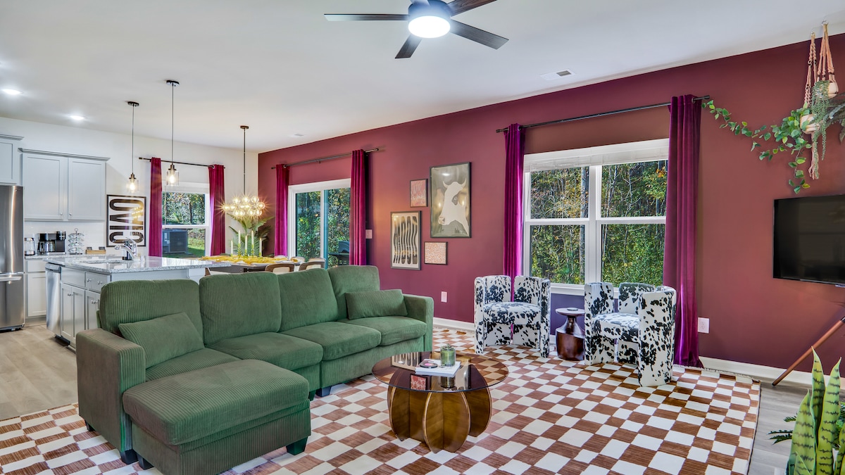 The living room offers a spacious layout with a plush green couch and two patterned chairs. Large windows are framed by vibrant curtains, welcoming natural light. A modern coffee table centers the room, and the decor features artistic elements on the walls.